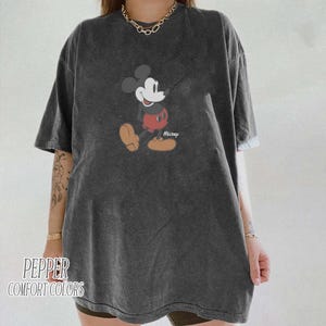 Op de afbeelding: Donkergrijs T-shirt met een klassieke Mickey Mouse afbeelding. Het stripfiguur is afgebeeld in zijn iconische pose, met een rode broek en gele schoenen. De tekst "Mickey" staat onder het personage. Het shirt heeft ook de tekst "Pepper Comfort Colors".