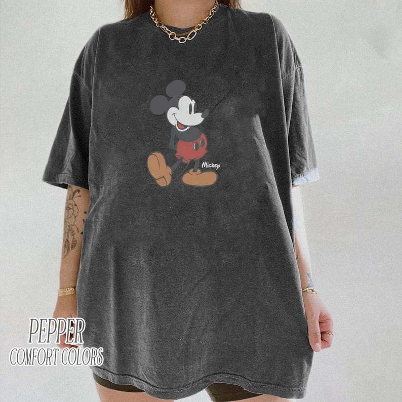 Op de afbeelding: Donkergrijs T-shirt met een klassieke Mickey Mouse afbeelding. Het stripfiguur is afgebeeld in zijn iconische pose, met een rode broek en gele schoenen. De tekst "Mickey" staat onder het personage. Het shirt heeft ook de tekst "Pepper Comfort Colors".