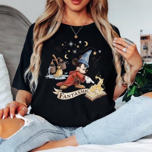 May include: Black t-shirt featuring a cartoon Mickey Mouse dressed as a sorcerer, with a red robe and blue hat. The design includes stars, moons, and the word "FANTASMIC" on a banner. A whimsical design.