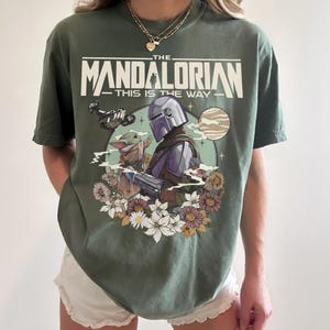 May include: Olive green t-shirt featuring a graphic of The Mandalorian and Grogu surrounded by flowers. The text "THE MANDALORIAN - THIS IS THE WAY" is printed above the characters. The design includes a spaceship and a planet.