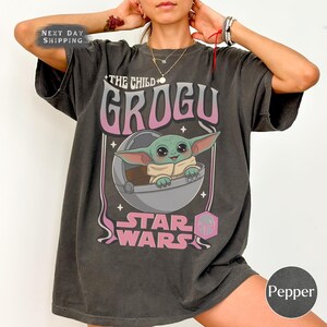 Puede incluir: Una camiseta de manga corta gris carb&oacute;n con un estampado gr&aacute;fico del personaje Grogu de Star Wars. El dise&ntilde;o incluye el texto THE CHILD GROGU y STAR WARS en letras rosas y blancas, con Grogu dentro de una c&aacute;psula circular.