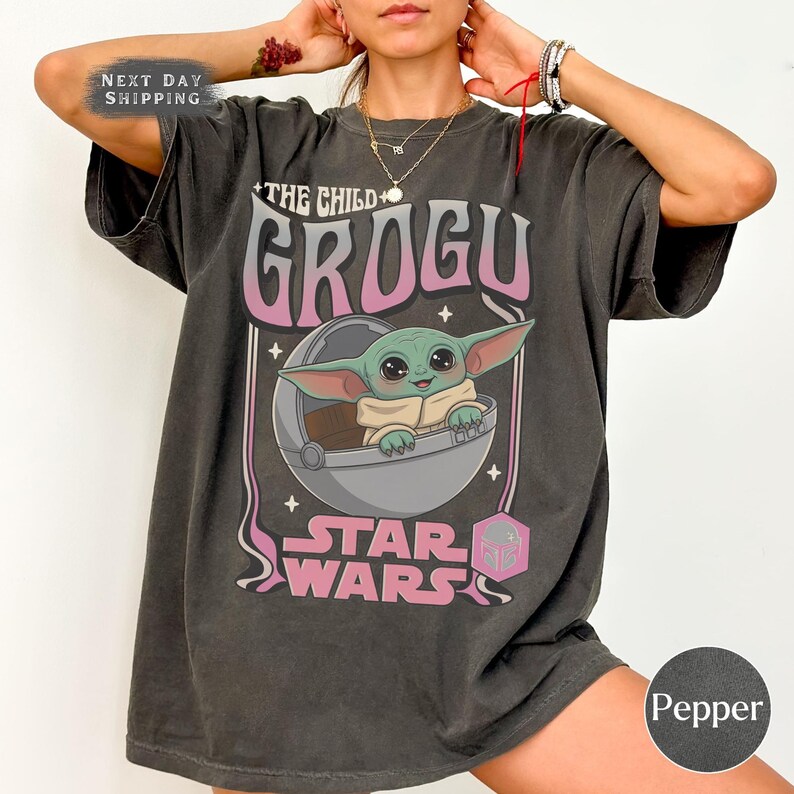 Puede incluir: Una camiseta de manga corta gris carb&oacute;n con un estampado gr&aacute;fico del personaje Grogu de Star Wars. El dise&ntilde;o incluye el texto THE CHILD GROGU y STAR WARS en letras rosas y blancas, con Grogu dentro de una c&aacute;psula circular.
