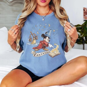 May include: Light blue t-shirt featuring a cartoon Mickey Mouse dressed as a sorcerer. Mickey is wearing a red robe and a blue wizard hat, with the word "Fantasmic" on a banner below. The design includes stars, moons, and a magic book.