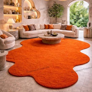 May include: An orange, irregularly shaped rug dominates the room's floor. The rug is placed in front of a cream-colored sectional sofa with orange and brown accent pillows. The room has neutral-toned walls, decorative shelves, and pendant lights.