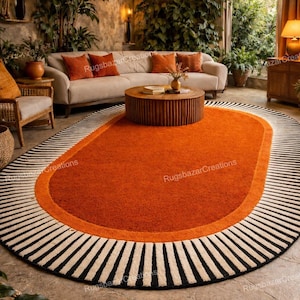 May include: An oval-shaped rug with a vibrant orange center, bordered by a lighter orange band and a black and white striped edge. The rug is placed in a living room setting with a neutral-toned sofa and decorative pillows.