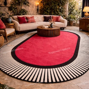 May include: Oval red rug with a black border and a white and black striped design. The rug is in a living room setting with a cream-colored sofa, a wooden coffee table, and hanging pendant lights.