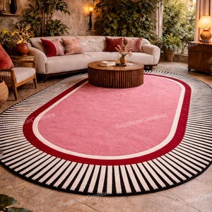 May include: Oval pink rug with a red and white border, and a black and white striped outer edge. The rug is in a living room setting with a white sofa, wooden coffee table, and hanging pendant lights.
