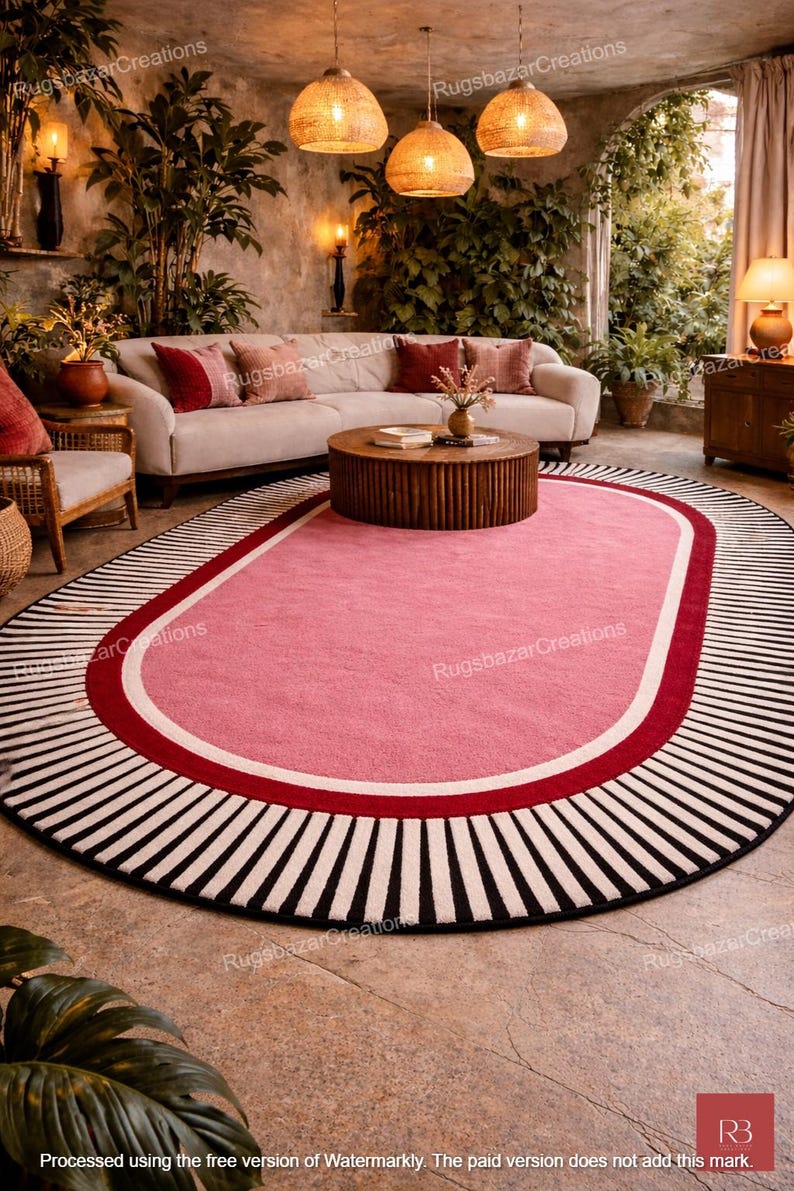 May include: Oval pink rug with a red and white border, and a black and white striped outer edge. The rug is in a living room setting with a white sofa, wooden coffee table, and hanging pendant lights.