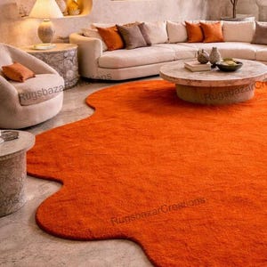 May include: An orange, irregularly shaped area rug dominates the foreground of a living room scene. The rug's vibrant color contrasts with the neutral tones of the furniture and walls. A curved sofa, armchairs, and a coffee table are also visible.