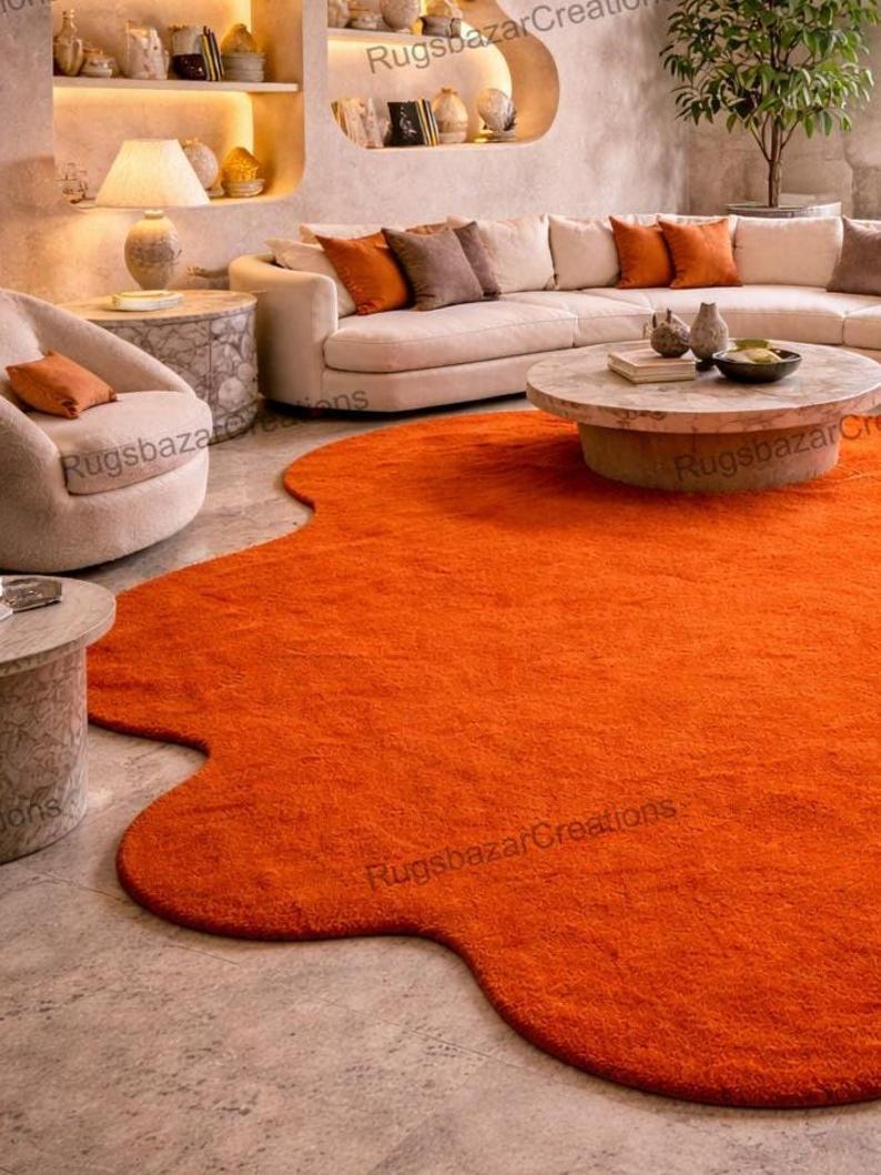 May include: An orange, irregularly shaped area rug dominates the foreground of a living room scene. The rug's vibrant color contrasts with the neutral tones of the furniture and walls. A curved sofa, armchairs, and a coffee table are also visible.