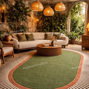 May include: An oval green rug with a rust-colored border and a striped outer edge. The rug is in a living room setting with a beige sofa, wooden coffee table, and woven pendant lights. The room has a natural, earthy aesthetic.
