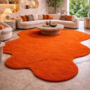 May include: An orange, irregularly shaped area rug dominates the room's center. The rug's plush texture contrasts with the neutral-toned furniture, including a curved sofa with orange and brown accent pillows, a round coffee table, and a side chair.