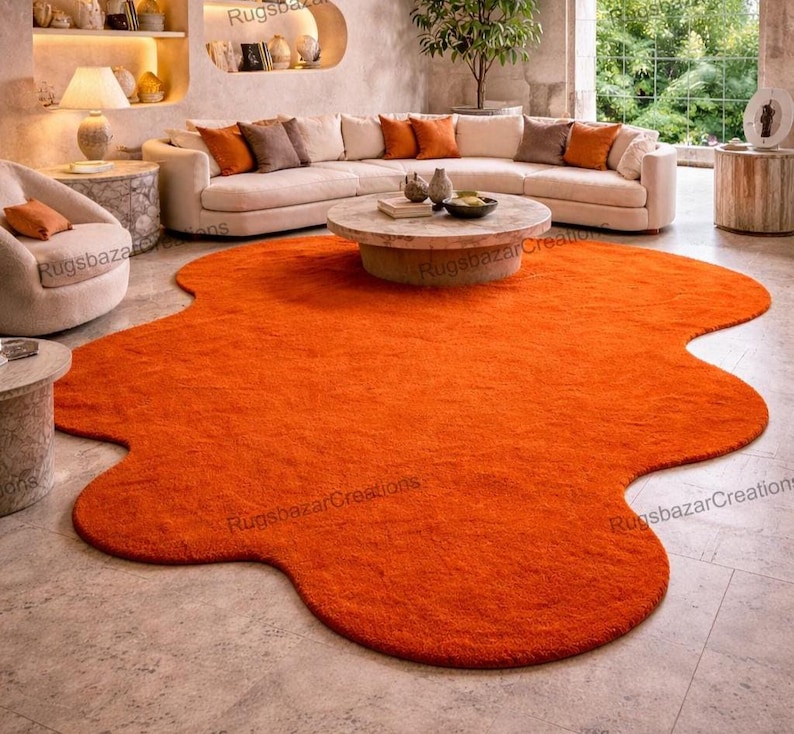 May include: An orange, irregularly shaped area rug dominates the room's center. The rug's plush texture contrasts with the neutral-toned furniture, including a curved sofa with orange and brown accent pillows, a round coffee table, and a side chair.