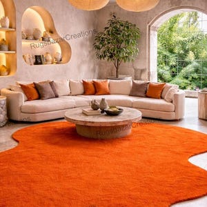 May include: An orange, irregularly shaped rug dominates the foreground of a living room scene. A beige sectional sofa with orange and brown throw pillows is in the background. A round, light-colored coffee table sits on the rug. The room has a neutral color palette.