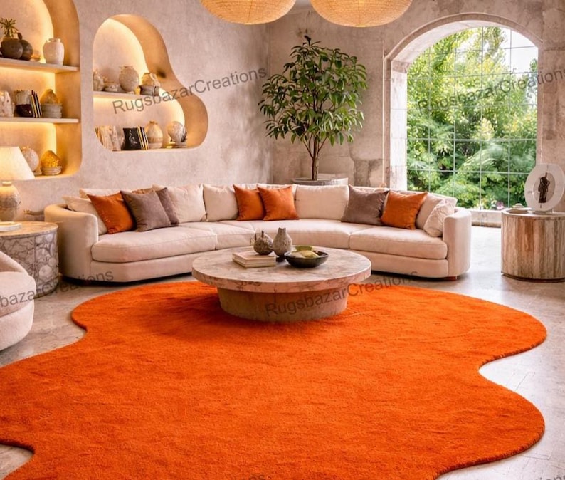 May include: An orange, irregularly shaped rug dominates the foreground of a living room scene. A beige sectional sofa with orange and brown throw pillows is in the background. A round, light-colored coffee table sits on the rug. The room has a neutral color palette.