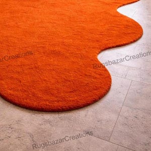 May include: An orange, irregularly shaped rug with a textured surface. The rug is placed on a light gray tiled floor. The rug's edges are rounded and the overall shape is organic. The rug is a vibrant accent piece.