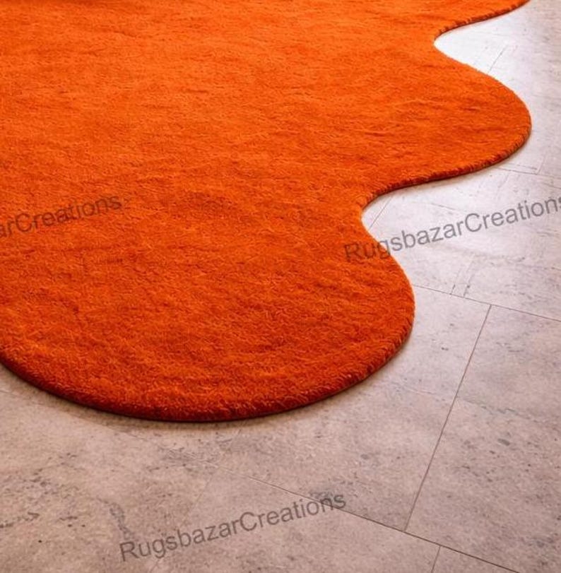 May include: An orange, irregularly shaped rug with a textured surface. The rug is placed on a light gray tiled floor. The rug's edges are rounded and the overall shape is organic. The rug is a vibrant accent piece.