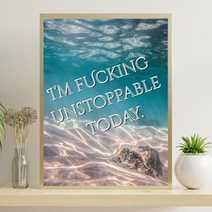 May include: A rectangular framed wall art print featuring an underwater scene with sunlight filtering through clear blue water onto a sandy bottom with a rock. The text I'M FUCKING UNSTOPPABLE TODAY is printed in white capital letters across the center.