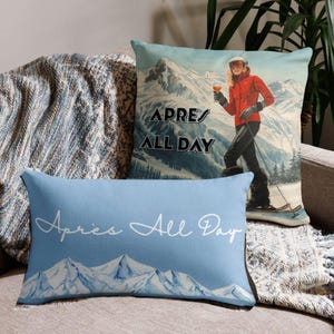 Könnte beinhalten: Ein quadratisches Kissen mit einem Skifahrer in roter Jacke, der ein Getränk hält, mit dem Text APRES ALL DAY, und ein rechteckiges hellblaues Kissen mit weißer Bergillustration und dem Schriftzug Apres All Day.