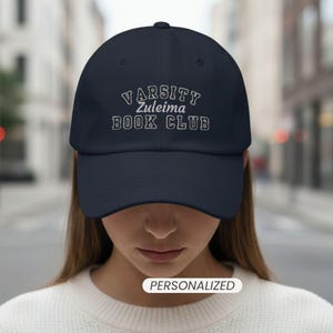 May include: A navy blue baseball cap featuring white embroidered text that reads VARSITY Zuleima BOOK CLUB. The cap has a curved brim and a structured crown. A small white rectangular label with the word PERSONALIZED is visible below.