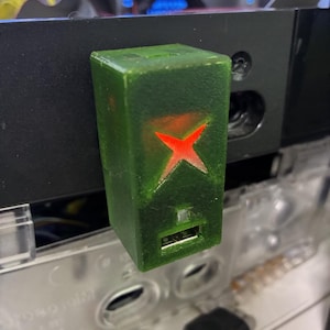 May include: A green USB adapter with a red Xbox logo, attached to a black electronic device. The adapter has a USB port. Other electronic components are visible in the background. The adapter is rectangular.