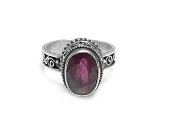 Op de afbeelding: Een zilveren ring met een ovale, dieproze edelsteen. De ring heeft een sierlijk ontwerp rond de edelsteen en op de band, met kleine kralen en wervelende patronen. De edelsteen is het middelpunt, gezet in een gedetailleerde zilveren zetting.