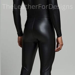 May include: A black, form-fitting jumpsuit with long sleeves and a high neckline. The LeatherForDesigns logo is visible. The jumpsuit appears to be made of a shiny, leather-like material. The model is facing away from the camera.