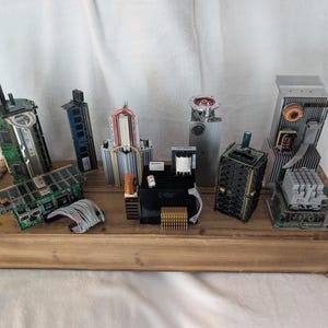 Pode incluir: Uma coleção de componentes de computador reaproveitados dispostos em uma bandeja de madeira. Os componentes incluem placas de circuito, dissipadores de calor e outras peças eletrônicas, montados em formas abstratas.