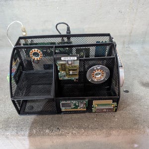 Puede incluir: Un organizador de escritorio de malla de alambre negro reutilizado con componentes de computadora. El organizador tiene múltiples compartimentos y contiene placas de circuito, un ventilador y otras piezas electrónicas. El diseño general es industrial y único.