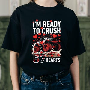 May include: Black t-shirt with a graphic featuring a monster truck design. The text reads "I'M READY TO CRUSH" above the truck and "67 HEARTS" below. Red hearts are scattered around the truck and text.