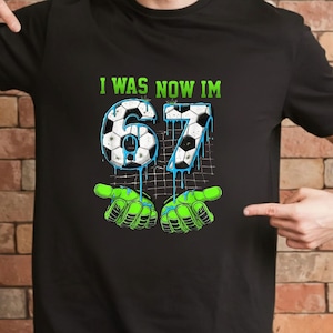 May include: Black t-shirt with the text "I WAS NOW IM" above the number "67" designed as soccer balls with blue dripping accents. The design includes green goalie gloves.