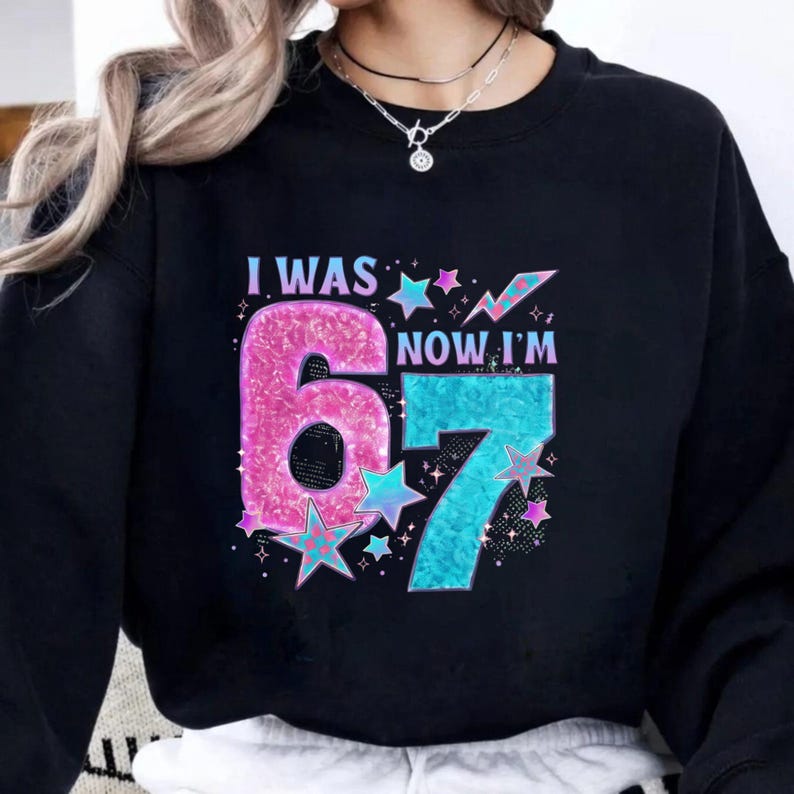 May include: Black sweatshirt with the text "I WAS 6 NOW I'M 7" in pink and blue glittery font. The number 6 is pink, and the number 7 is blue. Stars and sparkles surround the numbers.