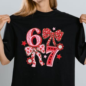 May include: Black t-shirt featuring the numbers "67" in a decorative design. The numbers are in pink and red with bow and floral accents. The design includes stars and hearts.