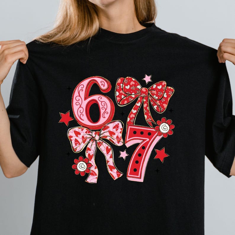 May include: Black t-shirt featuring the numbers "67" in a decorative design. The numbers are in pink and red with bow and floral accents. The design includes stars and hearts.