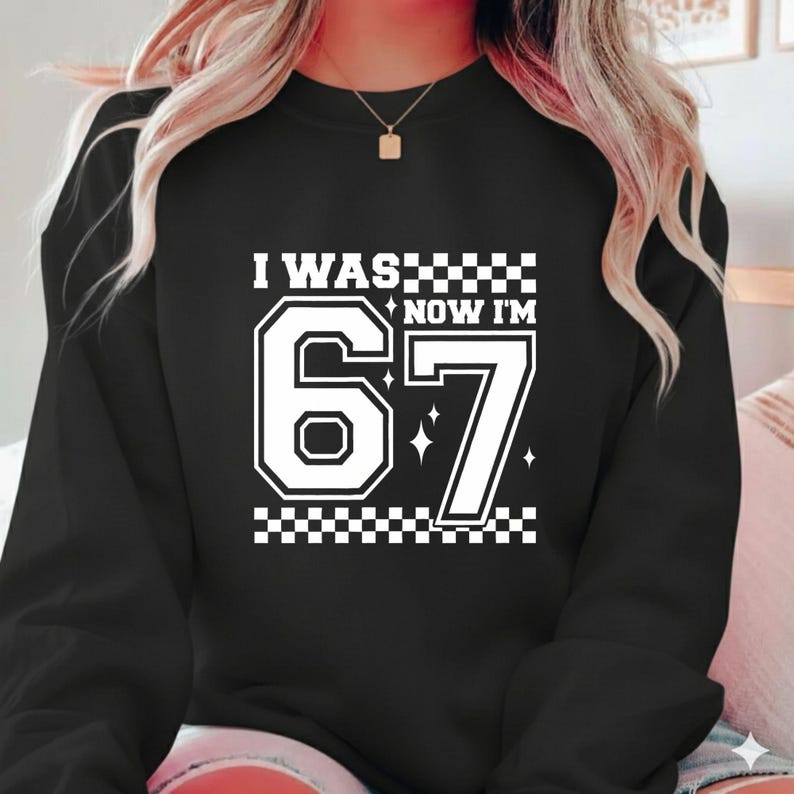 May include: Black sweatshirt with the text "I WAS... NOW I'M 67" in white. The number 67 is large and outlined in white, with a checkerboard pattern above and below the text. The sweatshirt has a crew neck.