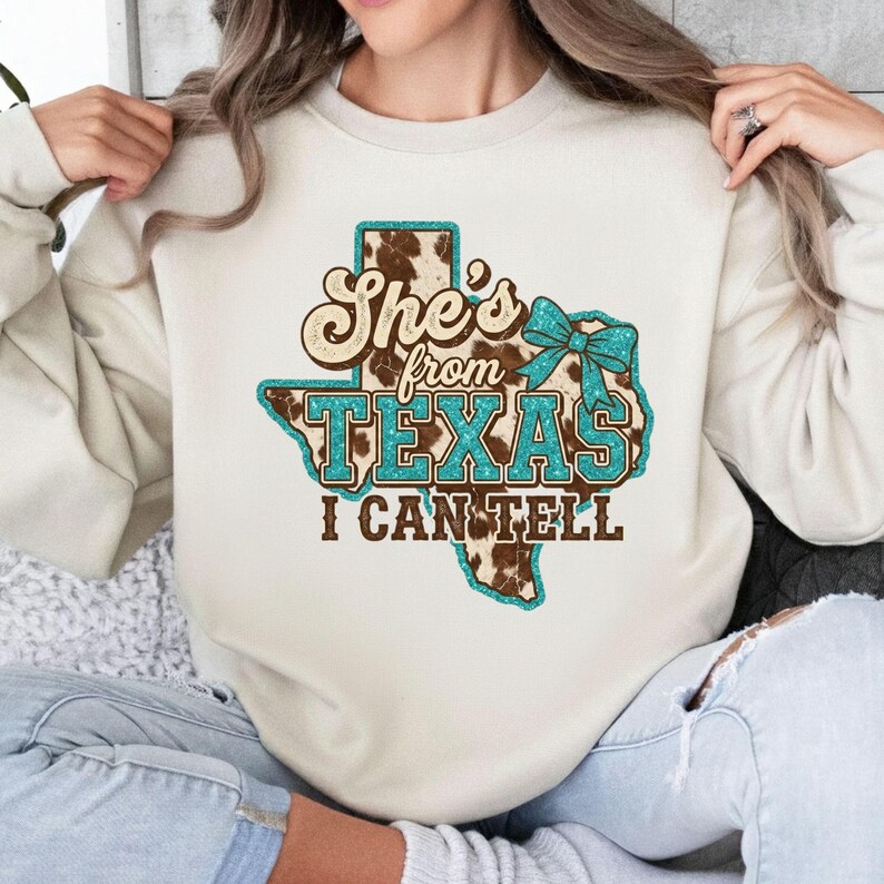 May include: A cream-colored sweatshirt with a graphic design featuring the state of Texas. The design includes the text "She's from TEXAS I CAN TELL" in brown and turquoise, with a cowhide pattern and a turquoise bow.