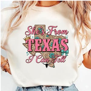 May include: A cream-colored t-shirt featuring a pink and brown graphic of the state of Texas. The graphic includes the text "She's From Texas I Can Tell" with turquoise accents and a longhorn silhouette.