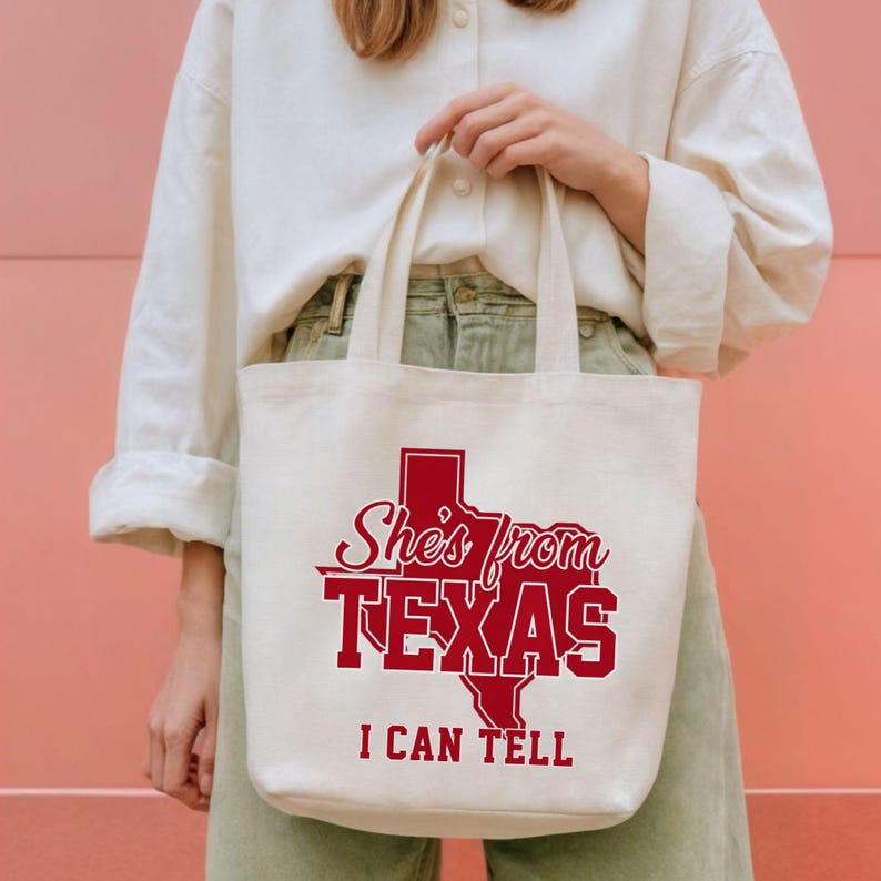 May include: Cream-colored tote bag with red text and a Texas outline. The text reads "She's from TEXAS I CAN TELL". The bag has two handles and is held by a person.