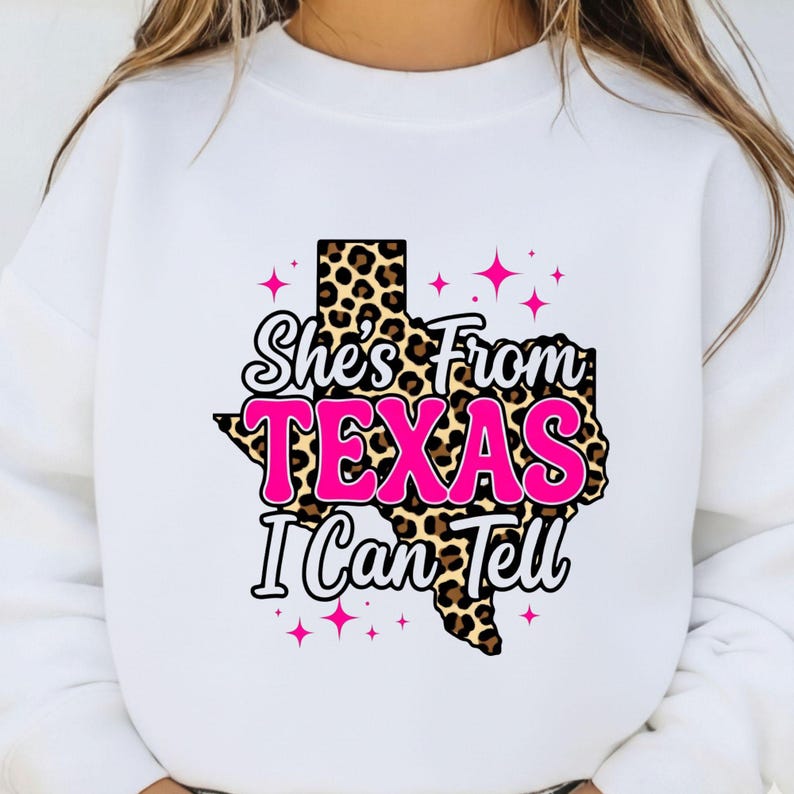 May include: White sweatshirt featuring a graphic of the state of Texas in a leopard print design. The text "She's From TEXAS I Can Tell" is in a mix of pink and white fonts, with pink star accents.