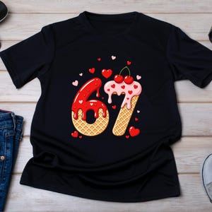 May include: Black t-shirt featuring a whimsical design of the number 67, crafted to resemble an ice cream cone. The number 6 is red with dripping glaze, while the 7 is pink with cherries. Small red and pink hearts are scattered around the numbers.