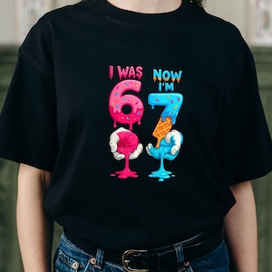 May include: Black t-shirt with a colorful graphic that reads "I WAS 6 NOW I'M 7". The numbers are designed to look like melting ice cream cones. The number 6 is pink and the number 7 is blue.