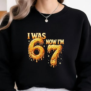 May include: Black sweatshirt with the text "I WAS 67 NOW I'M" in gold and orange. The numbers 6 and 7 are designed to look like donuts with sprinkles and dripping glaze. The design includes star accents.