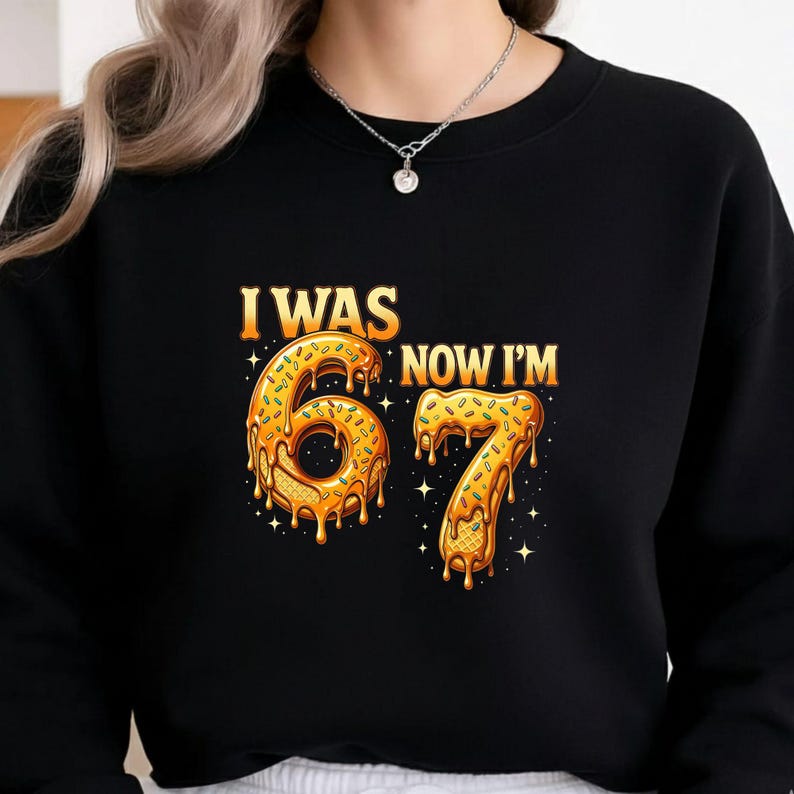 May include: Black sweatshirt with the text "I WAS 67 NOW I'M" in gold and orange. The numbers 6 and 7 are designed to look like donuts with sprinkles and dripping glaze. The design includes star accents.