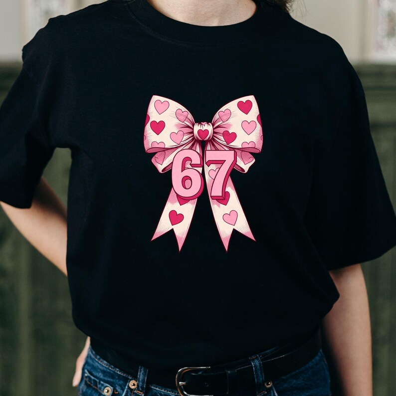 May include: Black t-shirt featuring a pink bow with heart patterns and the number 67 in pink. The bow has a light pink background with dark pink hearts. The number 67 is in a bold, pink font.