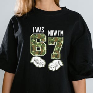 May include: Black t-shirt with the text "I WAS NOW I'M 67" in white. The numbers 6 and 7 are filled with a green and brown camouflage pattern. Two white open hands are below the numbers.