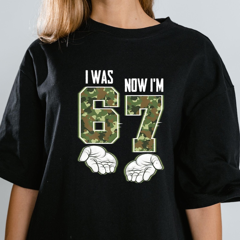 May include: Black t-shirt with the text "I WAS NOW I'M 67" in white. The numbers 6 and 7 are filled with a green and brown camouflage pattern. Two white open hands are below the numbers.