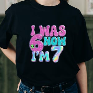 May include: Black t-shirt with the text "I WAS 6 NOW I'M 7" in pink, teal, and iridescent colors. The number 6 has an alien face. A fun birthday shirt for a child.