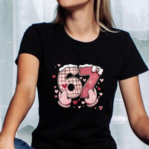 May include: Black t-shirt featuring the number "67" in a playful design. The "6" is a plaid pattern, and the "7" is a heart pattern, both with a white trim. Pink hearts and mittens surround the numbers, creating a festive look.
