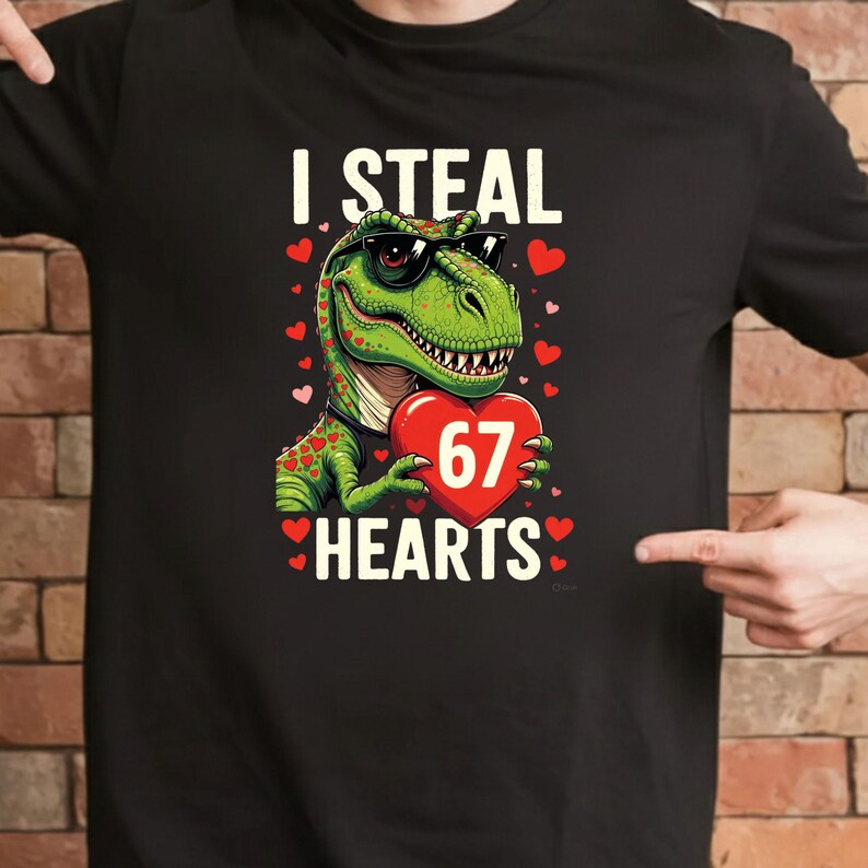 May include: Black t-shirt featuring a green dinosaur wearing sunglasses and holding a red heart with the number 67. The text "I STEAL 67 HEARTS" is printed above and below the dinosaur.