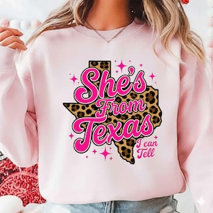 May include: A light pink sweatshirt with the text "She's From Texas I can Tell" in a pink and black design. The design includes a leopard print Texas outline and pink star accents.